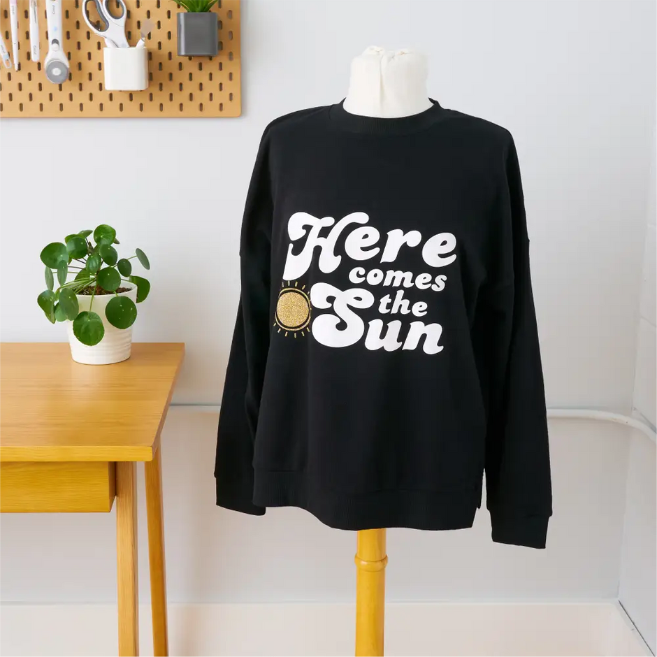 Black sweatshirt with ’Here comes the Sun’ text and a sun graphic in white and yellow.